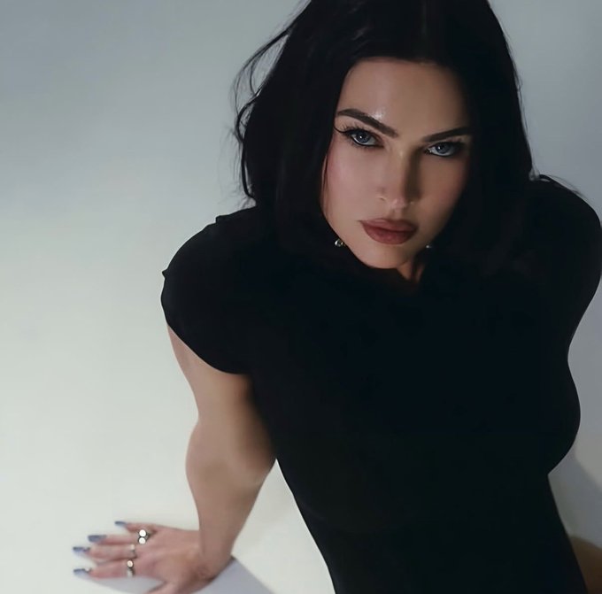 A glamorous portrait of a woman with dark hair, blue eyes, and a black top, posing against a light background with her hand on the surface, wearing rings and sporting dark nail polish.
