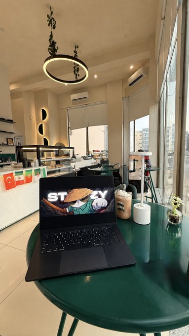 A laptop displaying a screensaver with a ninja character and "STAY" text sits on a green café table beside an iced coffee, capturing a focused "GM WEB3" work session in a bright, modern coffee shop.