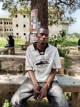 A young man sits on a bench wearing a white shirt featuring the "USDT" (Tether cryptocurrency) logo, with unfinished buildings and posters in the background—serving as a humorous visual reference to "USDT na the new skincare," suggesting the crypto logo looks like skincare branding.