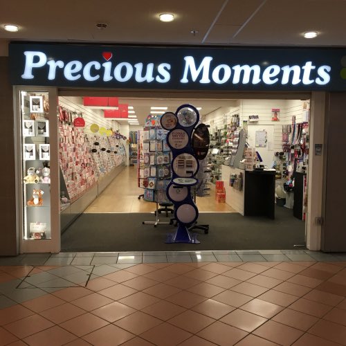 The image shows a store called "Precious Moments" with a display window featuring various items for sale. The store has a tiled floor and a sign that reads "Precious Moments". There are several displays of cards, books, and other merchandise inside the store. A person is visible in the background, possibly browsing through the items or entering the store.