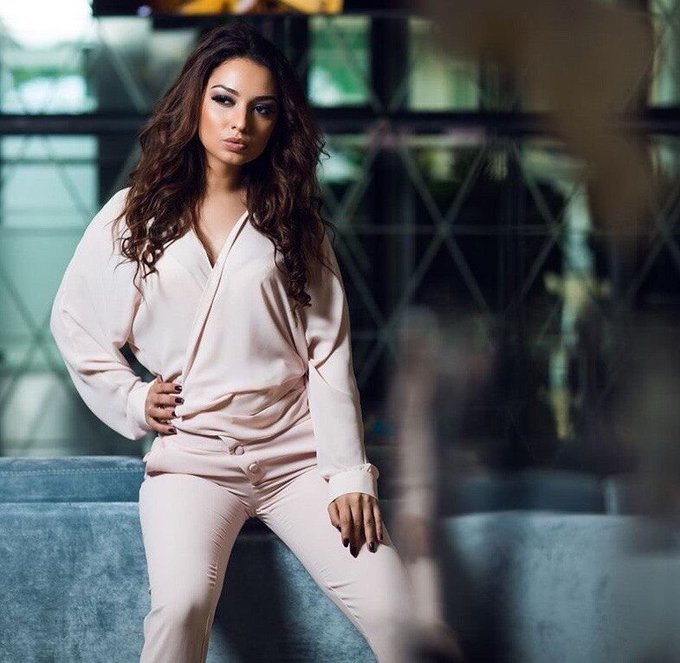 A portrait photograph of a woman with long, dark wavy hair posing in a light pink wrap-style blouse and matching pants. She sits on a blue velvet couch with a blurred, geometric background.