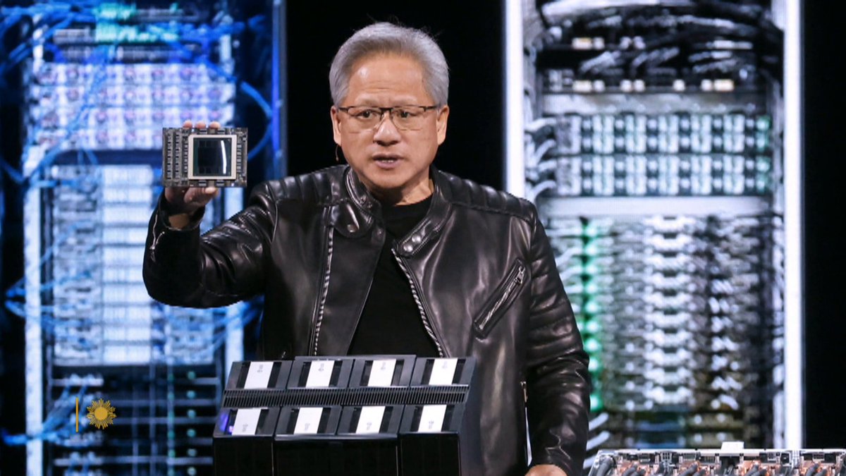 A man with gray hair, wearing a black leather jacket over a dark shirt, holds up a computer chip while speaking at a presentation, with server racks displaying blue-lit circuitry in the background. The scene appears to be a tech product launch or keynote event, fitting the context of a post by user "Hamm400."
