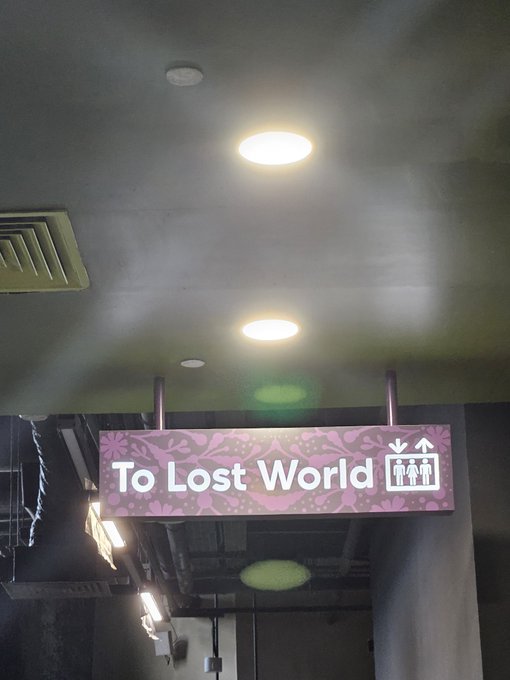 A pink "To Lost World" directional sign hangs from the ceiling, pointing visitors toward the attraction. The image suggests the start of an adventure, fitting the caption's optimistic tone of hoping to "win in the trenches."