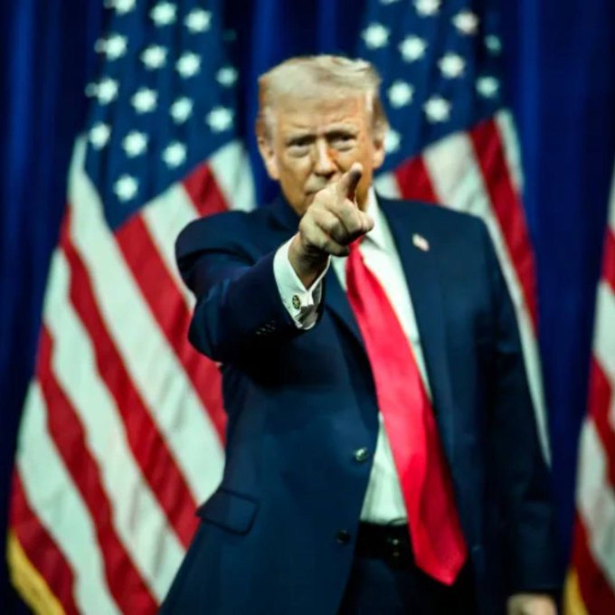 Donald Trump points assertively at the camera while standing before American flags, projecting a stern expression that mirrors his decisive stance in banning CNN, MS NOW, and Politico from the White House.