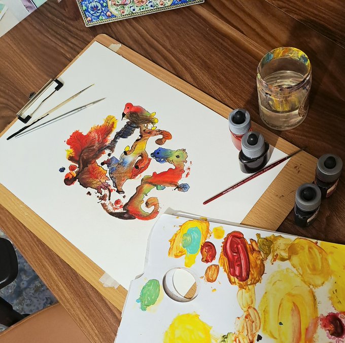 An artist's workspace featuring a vibrant watercolor and digitally collaged artwork titled "The Price of Silence" from the Collage Land series, depicting a fox with a red bird in a fluid, abstract composition of warm hues, alongside brushes, a palette with mixed paints, and bottles of ink on a wooden desk.