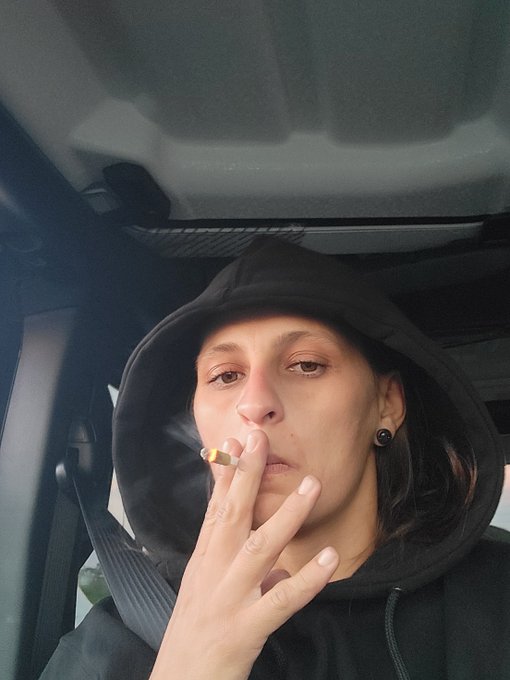A woman wearing a black hooded sweatshirt smokes a cigarette inside a car, embodying the casual, edgy aesthetic associated with the "Ladies of BAYC" (Bored Ape Yacht Club) community.