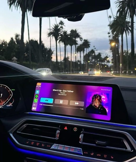 View from inside a car showing the infotainment screen playing 'Give It Up - Don Toliver' via SoundCloud, with palm trees and twilight visible through the windshield.