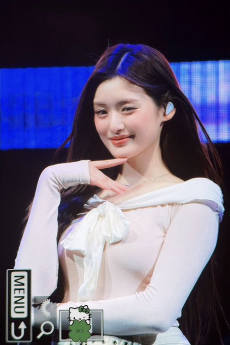 A young woman with long dark hair poses on stage wearing a white off-shoulder top, with a 'MENU' and Hello Kitty icon overlay visible on the photo.