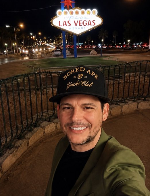 A man wearing a 'Bored Ape Yacht Club' cap taking a selfie in front of the illuminated 'Welcome to Fabulous Las Vegas Nevada' sign at night.