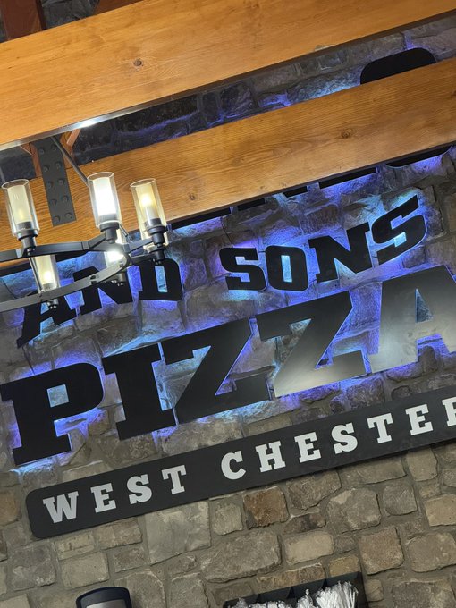An interior shot shows a metal sign reading 'AND SONS PIZZA WEST CHESTER' mounted on a stone wall beneath wooden ceiling beams and a chandelier.