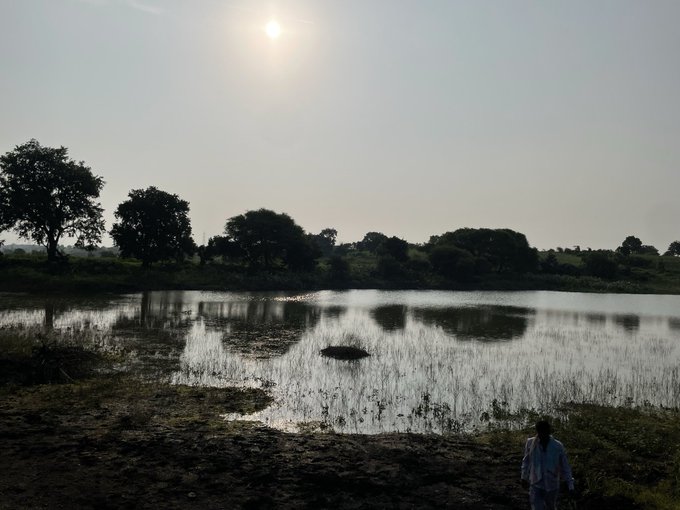 A person approaches a tranquil pond reflecting the hazy sun, with surrounding trees silhouetted against the pale sky—capturing the serene, immersive moment described in the "GMEN" post about enjoying a refreshing dip in nature.