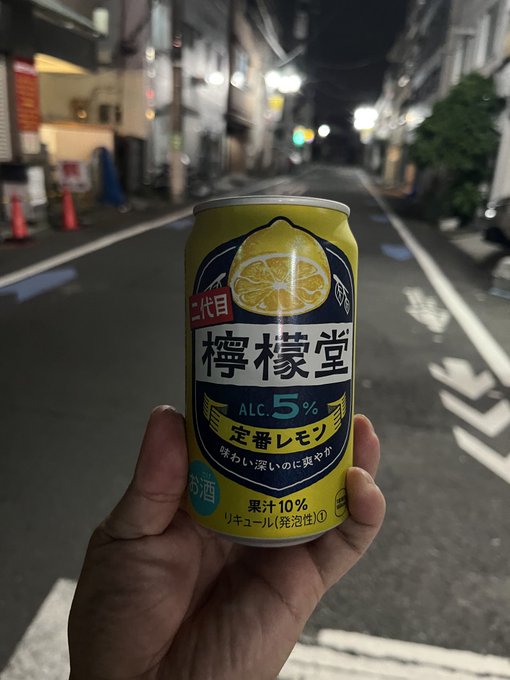 A photograph of a hand holding a yellow can of '檸檬堂' (Lemondo) lemon sour alcoholic drink with 5% alcohol on a Japanese street at night.