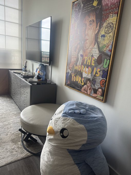 The image features a large stuffed penguin sitting on top of a white ottoman, with a poster hanging above it that says "The World Is Yours". The room also contains other items such as a television, a chair, and a window. There is a post on social media about the penguin plushie, which seems to be related to the image or the pudgypenguins series.
