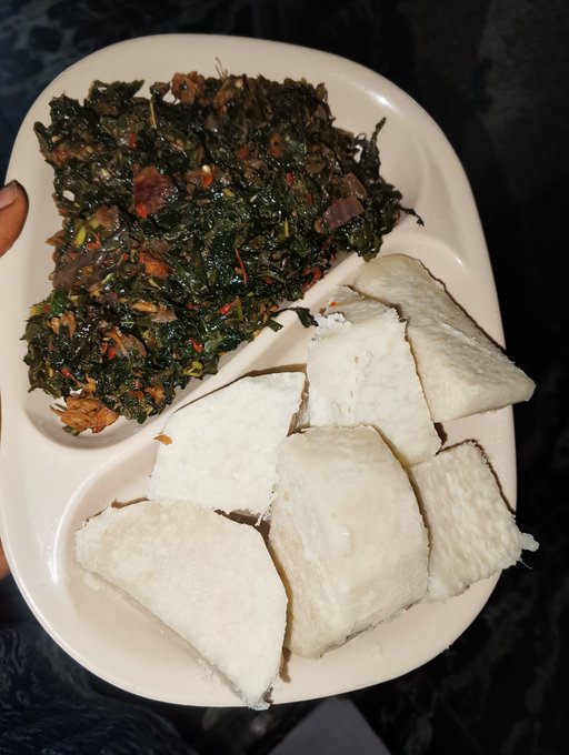 A white plate containing cooked greens with red peppers and onions, served alongside several pieces of a white starchy food, possibly cassava or yam.