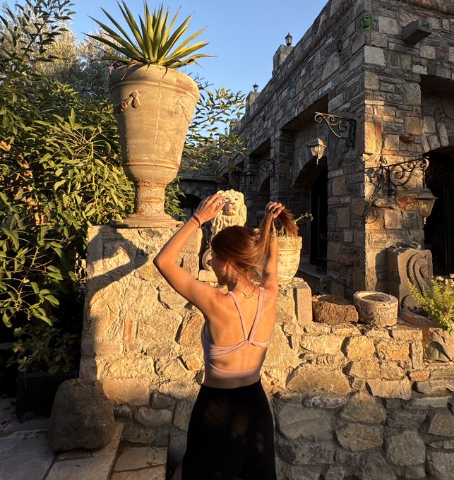A woman with her back to the camera poses with her arms raised in a stone courtyard featuring a large decorative urn and rustic architecture, bathed in warm golden sunlight. The image complements the caption's celebratory tone about a successful start to August, with the relaxed pose and scenic backdrop matching the post's grateful, optimistic mood about NFT collection performance.