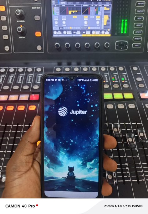 A hand holds a smartphone displaying the Jupiter crypto app logo on a cosmic wallpaper, set against an audio mixing console.