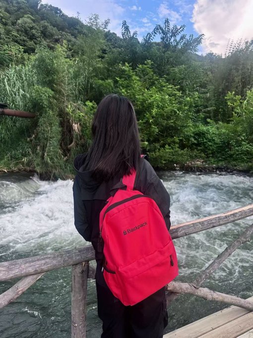 A person with long dark hair, wearing a black jacket and a red 'Basepack' backpack, stands at a wooden railing overlooking a rushing river surrounded by lush green vegetation.