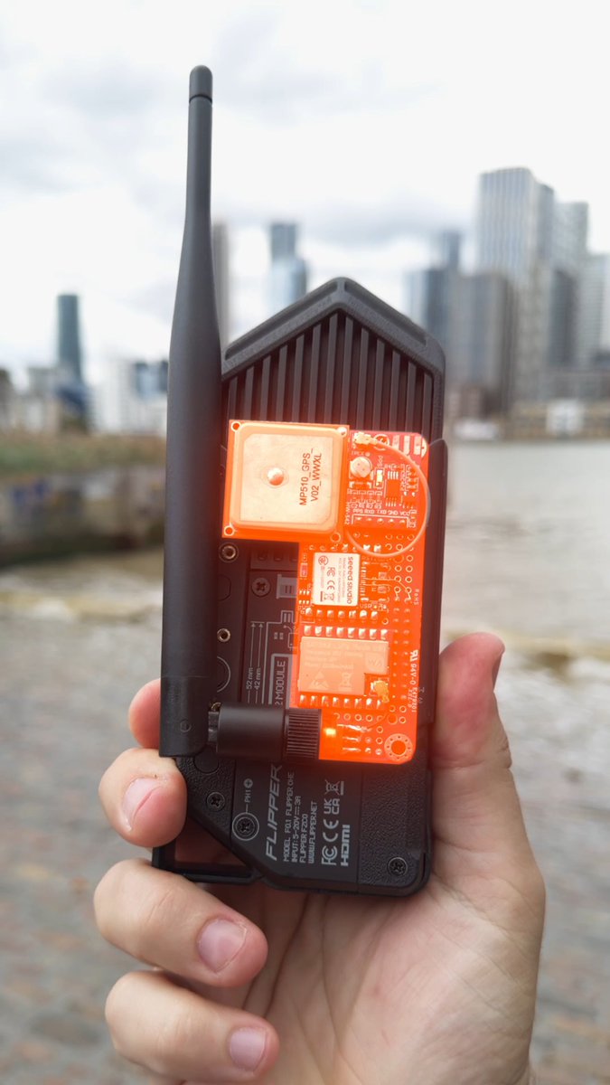A hand holds a custom black 'FLIPPER ONE' electronic device with an antenna and an exposed orange circuit board against a blurred city skyline and waterfront background.