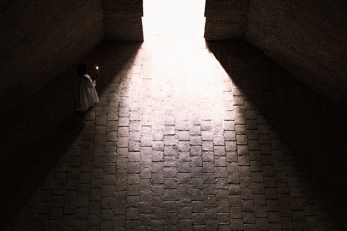 An atmospheric image of a small figure holding a candle, standing in a dark stone corridor illuminated by a triangular beam of light.