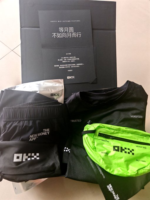An OKX promotional bundle labeled 'HAPPY MID-AUTUMN FESTIVAL 等月圆 不如问月而行' containing a black t-shirt with 'TRUSTED VERIFIED', shorts reading 'THE NEW MONEY App', and a neon green crossbody bag.