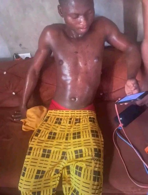 A shirtless young man wearing yellow patterned pants sits on a couch, drenched in sweat and holding a phone with charging cables attached.