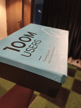 A blue box labeled '100M USERS' with smaller text reading 'Turning Choices into Decisions' sits on a dark surface, possibly a stool.