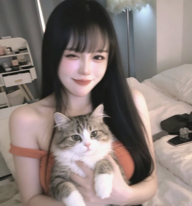 A young woman with long dark hair winks while holding a tabby cat, wearing an orange off-shoulder top in a bedroom setting.
