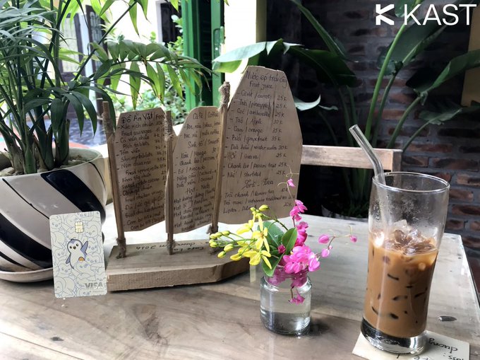 An iced coffee sits on a wooden café table alongside handwritten menu boards listing drinks like "Cà phê trứng" and "Egg coffee," with a small card featuring the KAST penguin mascot propped nearby, tying into the post's introduction of KAST as a global stablecoin-based financial platform during a solo Saturday morning coffee break.
