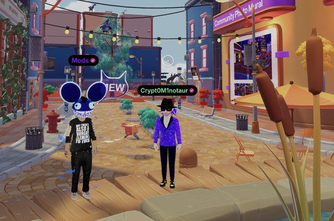 A screenshot from the Decentraland metaverse showing two avatars ('Mods' and 'CryptoMinotaur') standing in a vibrant virtual town square with the text 'Community Pte. to Meira!' visible.