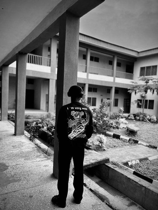 A black-and-white photo shows a person standing in a courtyard, viewed from behind, wearing a black jacket with a graphic that includes the text "I'm very well."