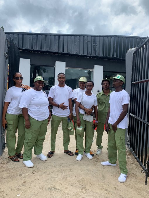 A group of eight young people wearing matching white t-shirts and olive green pants/caps pose together outside a modern dark-colored building.