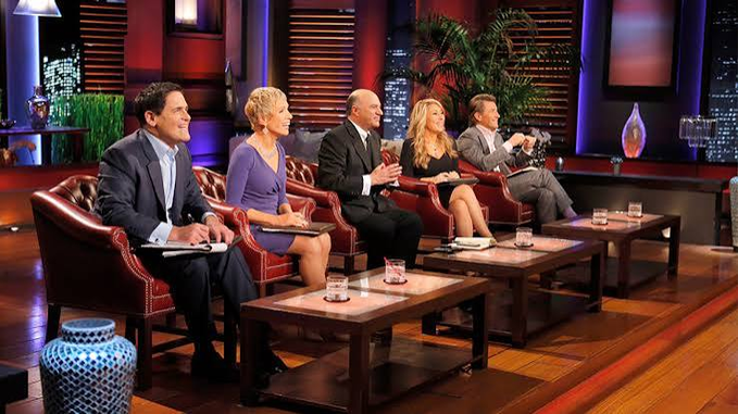 The cast of 'Shark Tank' sits in their signature leather chairs on the show's set, smiling and engaged in conversation.