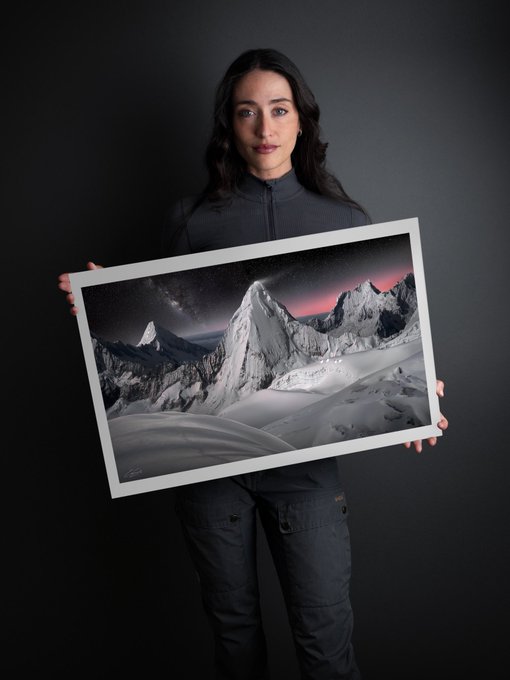 A woman with dark hair wearing a dark gray zip-up top and jeans holds a large framed landscape print depicting snowy mountain peaks against a starry pink sky.