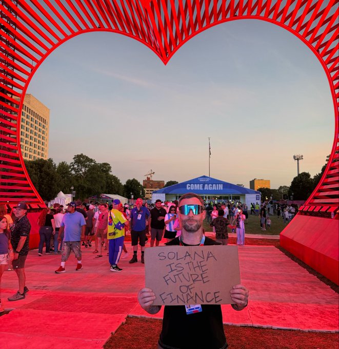 A man wearing reflective sunglasses stands under a large red heart sculpture, holding a cardboard sign that reads 'SOLANA IS THE FUTURE OF FINANCE'.