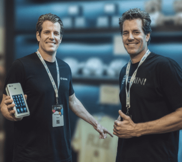 Two men smile while wearing black Gemini-branded t-shirts and lanyards, with one holding up a smartphone. The "Gemini" logo on their shirts reflects the post's context, as Cameron and Tyler Winklevoss co-founded Gemini, which is launching the Zcash ETF referenced in the caption.