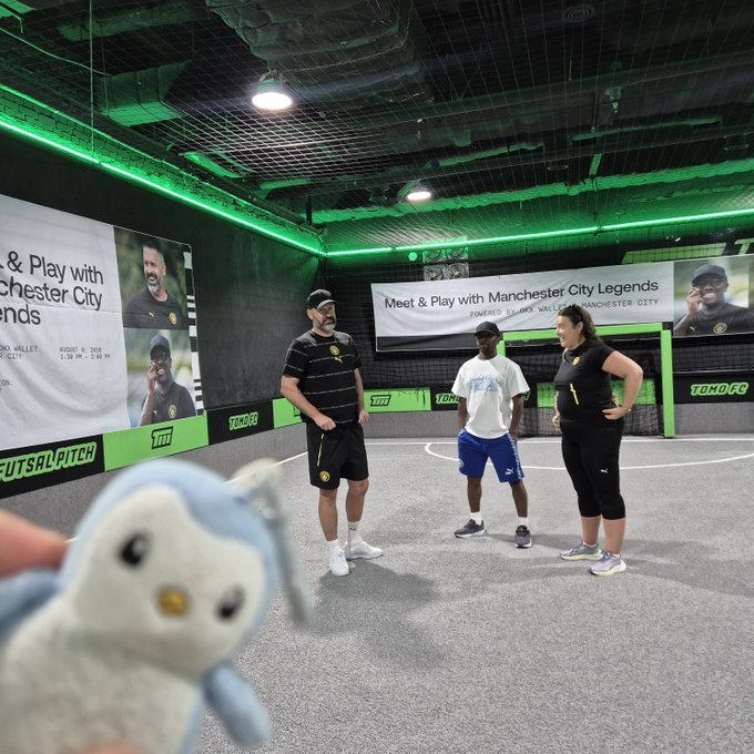 A photo taken at a 'TOMO' indoor soccer facility advertising 'Meet & Play with Manchester City Legends' (Roberto Di Matteo and Vincent Kompany), with a plush toy in the foreground.