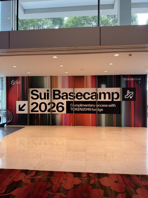 A large conference banner reading "Sui Basecamp 2026" with text "Complimentary access with TOKEN2049 badge" and the Sui logo and "TOKEN2049" branding displayed prominently in what appears to be an event venue lobby. The setup, two days before the event, marks the arrival of attendees and the imminent kickoff of the Sui-focused conference held alongside TOKEN2049.