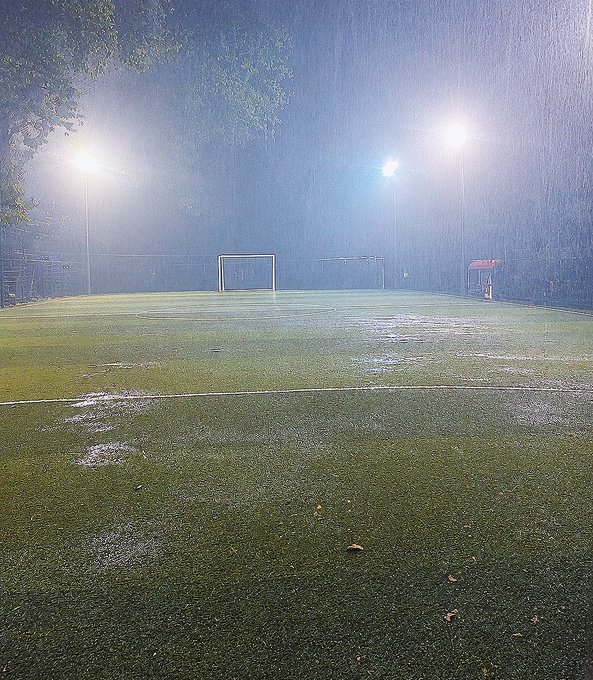 A nighttime photo of an empty soccer/football field illuminated by floodlights during heavy rain, showing water pooling on the artificial turf with a goalpost visible in the background.