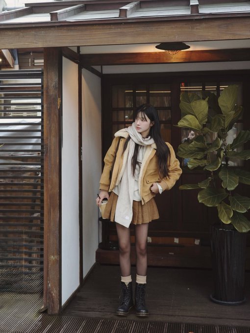 Woman wearing a tan jacket, large white scarf, pleated skirt, and black boots, standing on a wooden porch in front of a door, with a potted fiddle-leaf fig tree beside her.