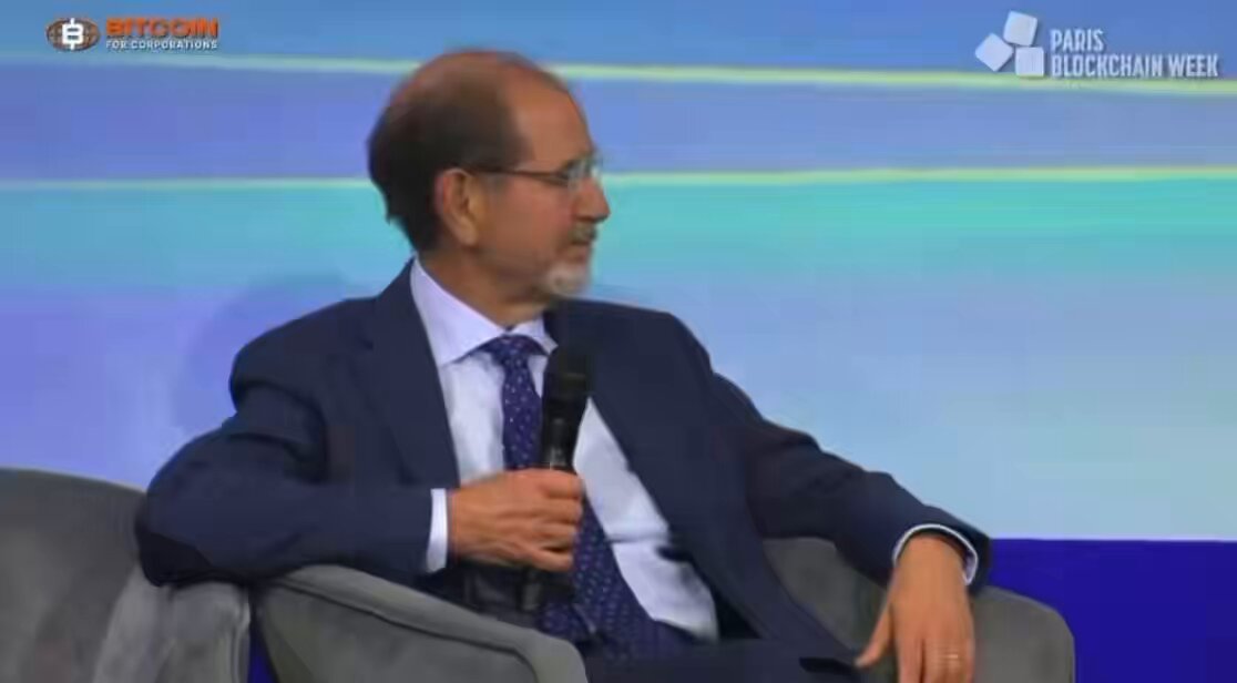 The image features a man sitting in a chair, wearing glasses and a suit jacket. He has his hand on a microphone, suggesting that he might be giving a speech or presentation. The background of the image shows a blue wall with white text, which appears to be related to the post about Bitcoin.