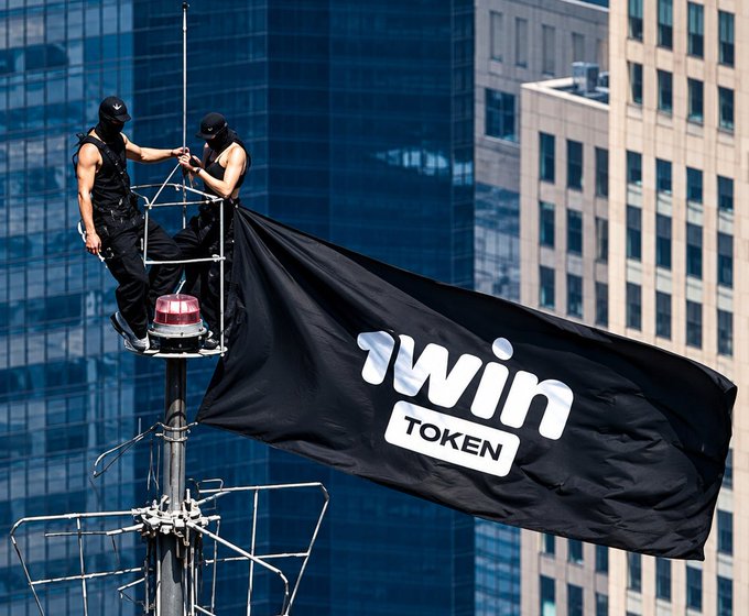 Two masked figures stand atop a communications tower flying a black flag bearing the white text '1WIN TOKEN' against a backdrop of glass skyscrapers.