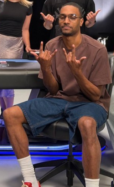 A man wearing a brown t-shirt, denim shorts, and glasses sits in a gaming-style chair making hand gestures, with another person in black making a rock-on sign behind him.