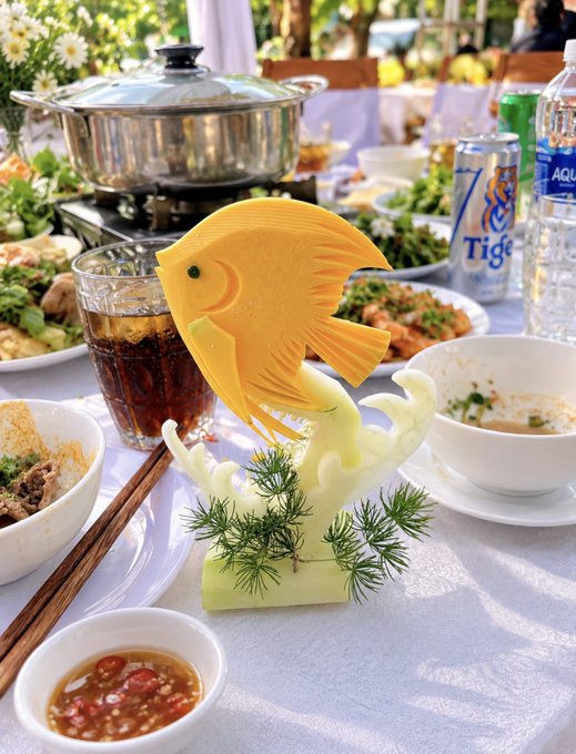 A beautifully carved mango sculpture shaped like a fish with green and white garnish base, displayed on a dining table surrounded by Vietnamese dishes, Tiger beer cans, and Aquafina water bottles.