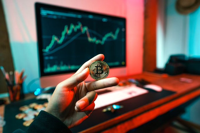 A person holds up a gold Bitcoin physical coin in front of a computer monitor displaying a green cryptocurrency trading chart, set in a room with moody red and blue lighting.
