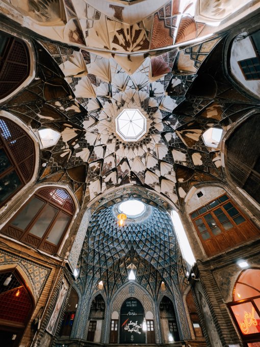 An interior photograph of an ornate historic bazaar or building (likely in Iran) showcasing intricate geometric tilework, stained glass windows, and a highly decorated dome with a central oculus.