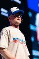 A man wearing a beige t-shirt, blue baseball cap, and sunglasses stands on a stage with a blue digital background.