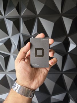 A hand holds up a sleek black "DCENT" biometric hardware wallet card against a geometric gray textured background, showcasing the device referenced in the post about securing crypto assets like $FLR.