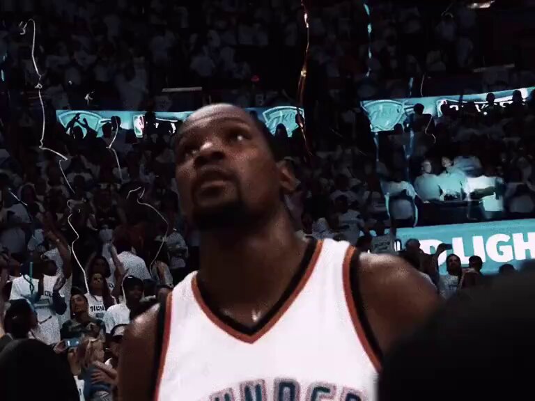 The image shows a basketball player wearing a white jersey with orange lettering, standing in front of a crowd at a basketball game. The player appears to be looking up into the air or towards something off-camera. There are several people around him, some sitting and others standing, all watching the game intently. A large screen is visible behind them, likely displaying the game's progress for everyone present. The image also contains text that reads "@rozikrypto is now officially part of @shadowcleague and things just got very big". This suggests that the basketball player might be a former professional athlete or has made a significant impact on the sport in some way.