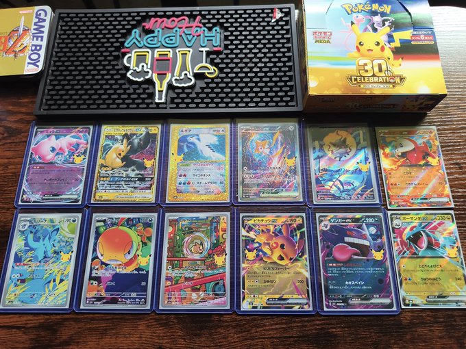 A collection of Pokémon cards is displayed on a wooden surface, including the Game Boy box and a Pokémon 30th Anniversary box.