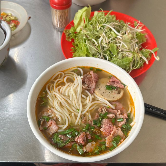 A bowl of Vietnamese noodle soup (appears to be bún bò Huế) with beef slices and chives sits on a metal table next to a red plate of fresh herbs and bean sprouts, with chopsticks bearing the handle '@0xdateth'.